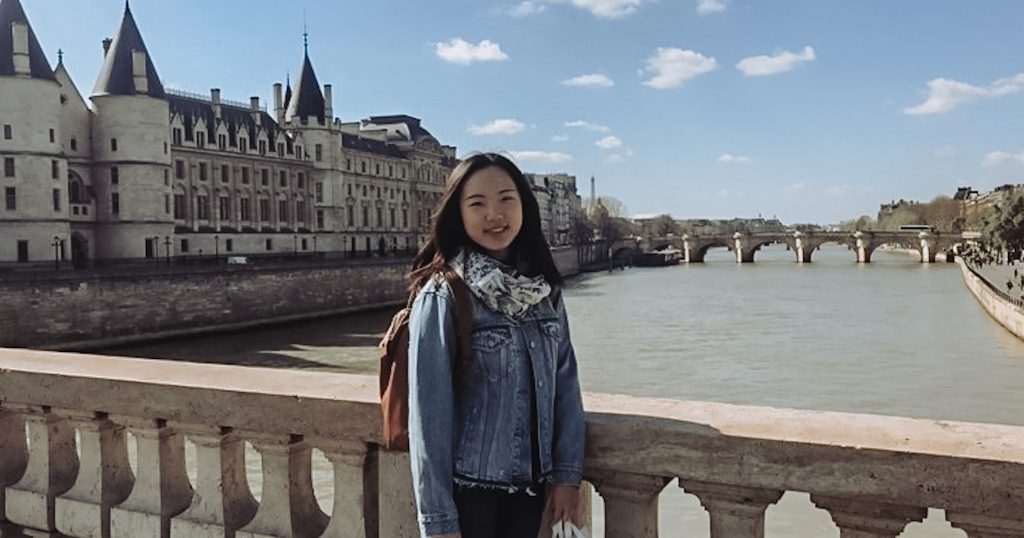 Hasel Chin, Gaststudentin aus China, auf einer Brücke. Im Hintergrund ein Schloss vor blauem Himmel.