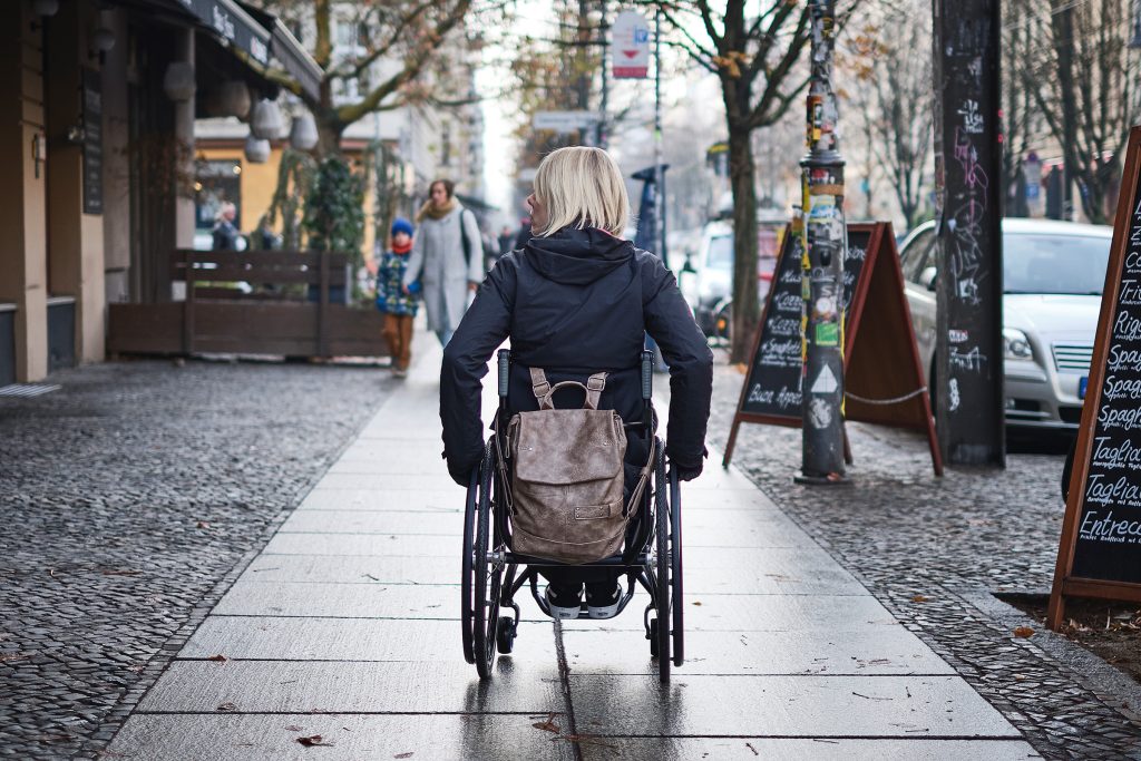 Foto einer Rollstuhlfahrerin, die in einer Stadt auf dem Gehweg entlangfährt.