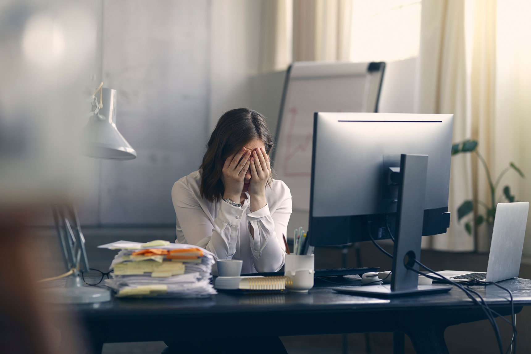 Foto einer Frau am Arbeitsplatz, die ihren Kopf in den Händen hält und gestresst wirkt. Auf ihrem Arbeitsplatz liegt ein grosser Stapel Papier mit Post-its, ein Block und Schreibutensilien, sowie ein Laptop und Bildschirm.