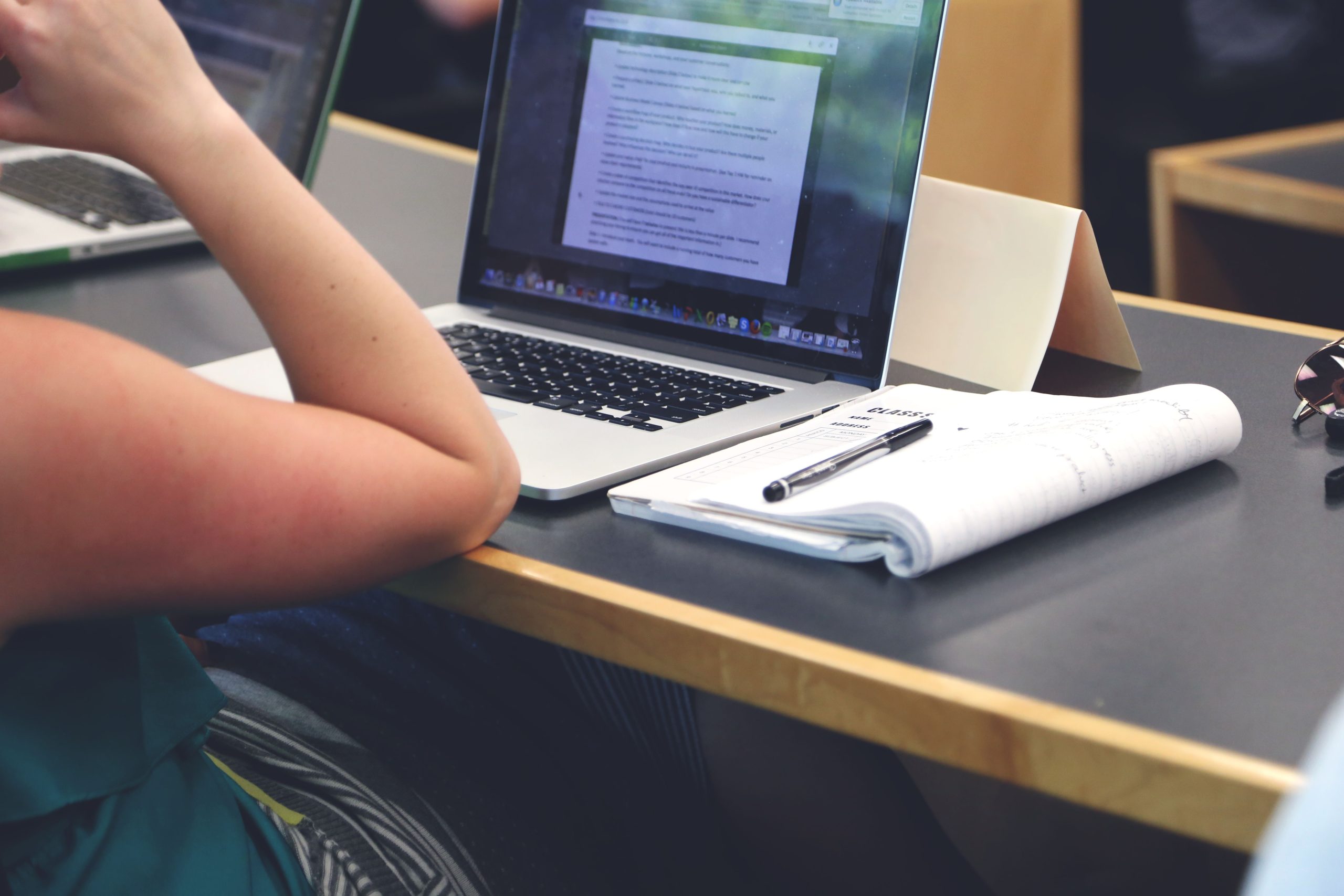 Person sitzt und arbeitet am Laptop; nur Arm der Person sichtbar. Notizblock und Stift liegen daneben bereit.