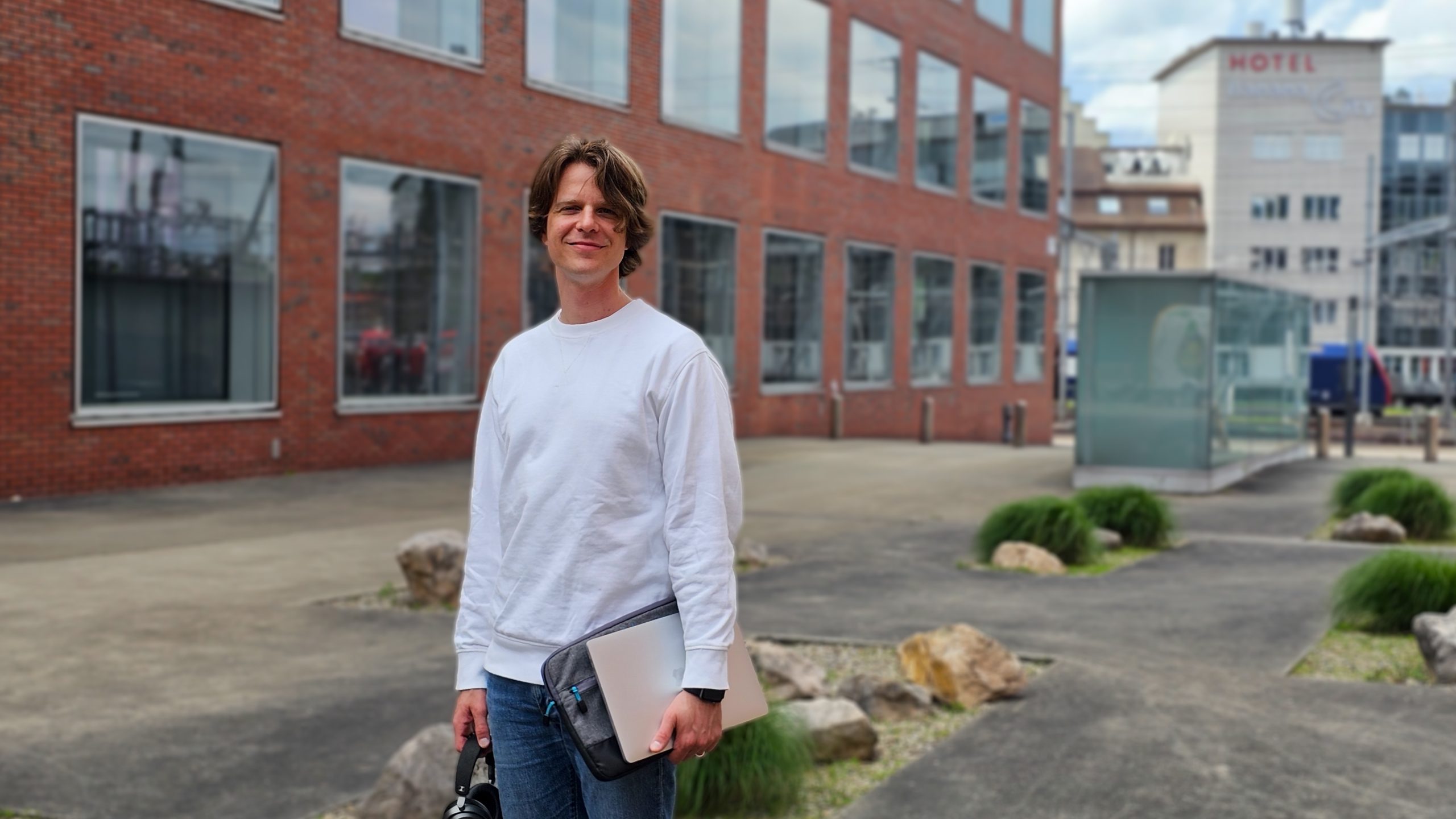 Auf dem Bild ist ein Mann mit einem weissem Pullover vor einem grossen Gebäude zu sehen, in der Hand trägt er einen PC. Hinter ihm sieht man den Bahnhof von Winterthur. Er macht gerade den Doktorat und erzählt von seinem PhD Programm