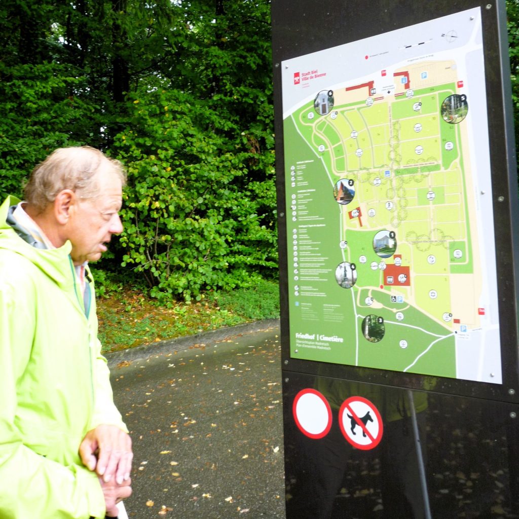 Ein älterer Mann betrachtet einen Übersichtsplan zur Orientierung auf dem Friedhof.
